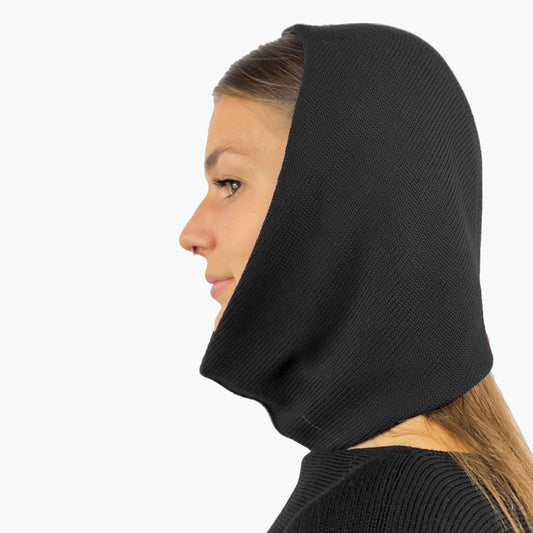 Eine junge Frau von der Seite zu sehen mit einem Nasenpiercing, trägt eine Mütze deren Form den Kopf umspielt, und auch das Kinn bedeckt, ähnlich einer geschlossenen Kapuze. Die Balaclava ist schwarz und aus Merinowolle gestrickt.