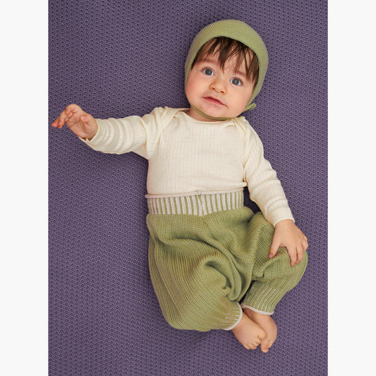 ein Baby mit einer Hellgrünen Haube liegt auf einer Lilafarbenen Babydecke und trägt eine Sarouelhose auch Harems- oder Pluderhose genannt. Mit tiefgeschnittenem Schritt und extrem viel Platz für Windeln. Die Matchagrüne Wollhose ist aus feinster Bio-Merinowolle gestrickt. Sie hat enganliegende hochelastischen Bündchen, die Wollweiße Längsstreifen haben.