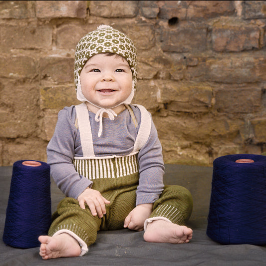 Lachendes Baby sitz zwischen Blauen Wollkonen und trägt eine gepunktete Mütze und die Woll-Hose, Salopette aus Bio-Merinowolle von vincente gestrickt. Sie ist Jeans-Blau mit Woll-weißen Hosenträgern und Woll-weißen Streifen im enganliegenden Bündchen. Die Knöpfe sind Beige und mit Roten Garn angenäht