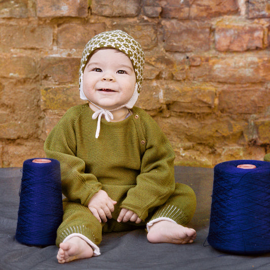 Lachendes Baby trägt eine gepunktete Mütze und eine Olivegrüne Babyjacke mit seitlicher Knöpfung, auch Schlüttli genannt. Die Strickjacke ist aus Bio-Merinowolle gestrickt