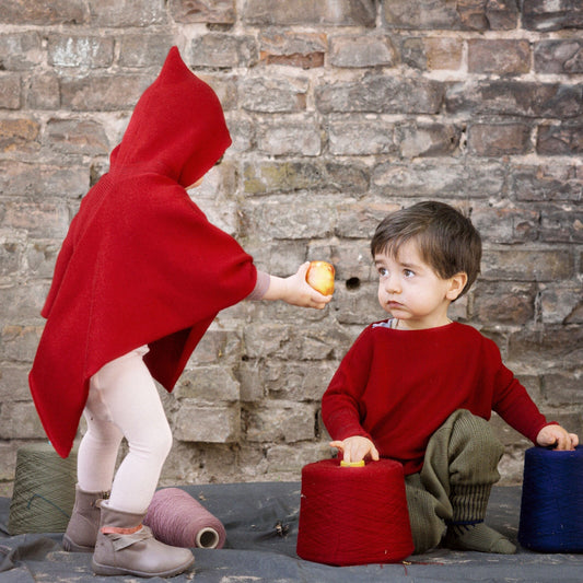 Ein kleines Mädchen mit einem roten Cape mit Kapuze reicht einem kritisch drein blickenden Jungen einen Apfel. Der Junge spielt lieber weiter mit den Wollkonen.