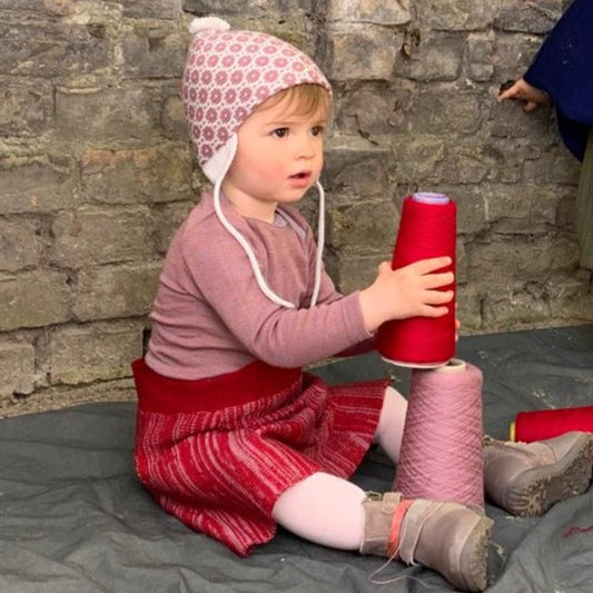 Ein kleines Mädchen baut einen Turm mit Wollkonen. Sie trägt eine Mütze von vincente und einen gestrickten Rock in Rosa und Rot.