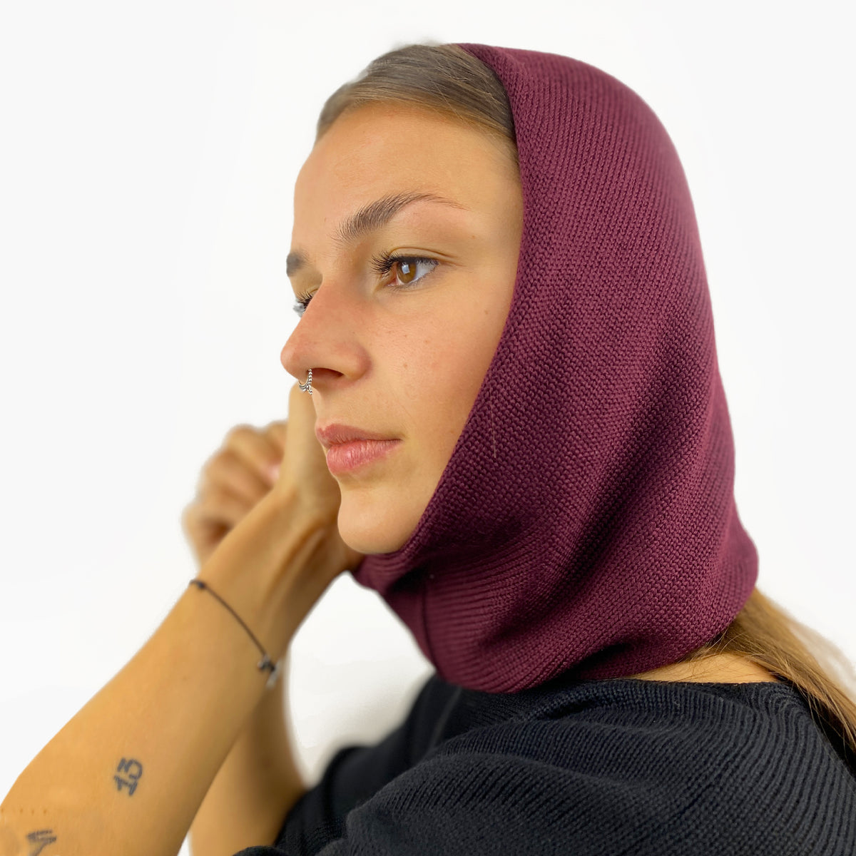 Eine junge Frau mit einem Nasenpiercing trägt eine Mütze deren Form den Kopf umspielt, und auch das Kinn bedeckt, ähnlich einer geschlossenen Kapuze. Die Balaclava ist Bordeauxrot und aus Merinowolle gestrickt.