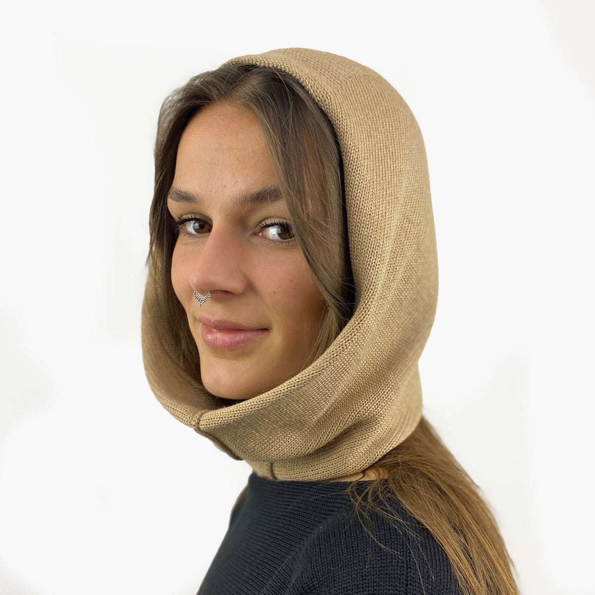 Eine junge Frau mit einem Nasenpiercing trägt eine Mütze deren Form den Kopf umspielt, und auch das Kinn bedeckt, ähnlich einer geschlossenen Kapuze. Die Balaclava ist Beige und aus Merinowolle gestrickt.