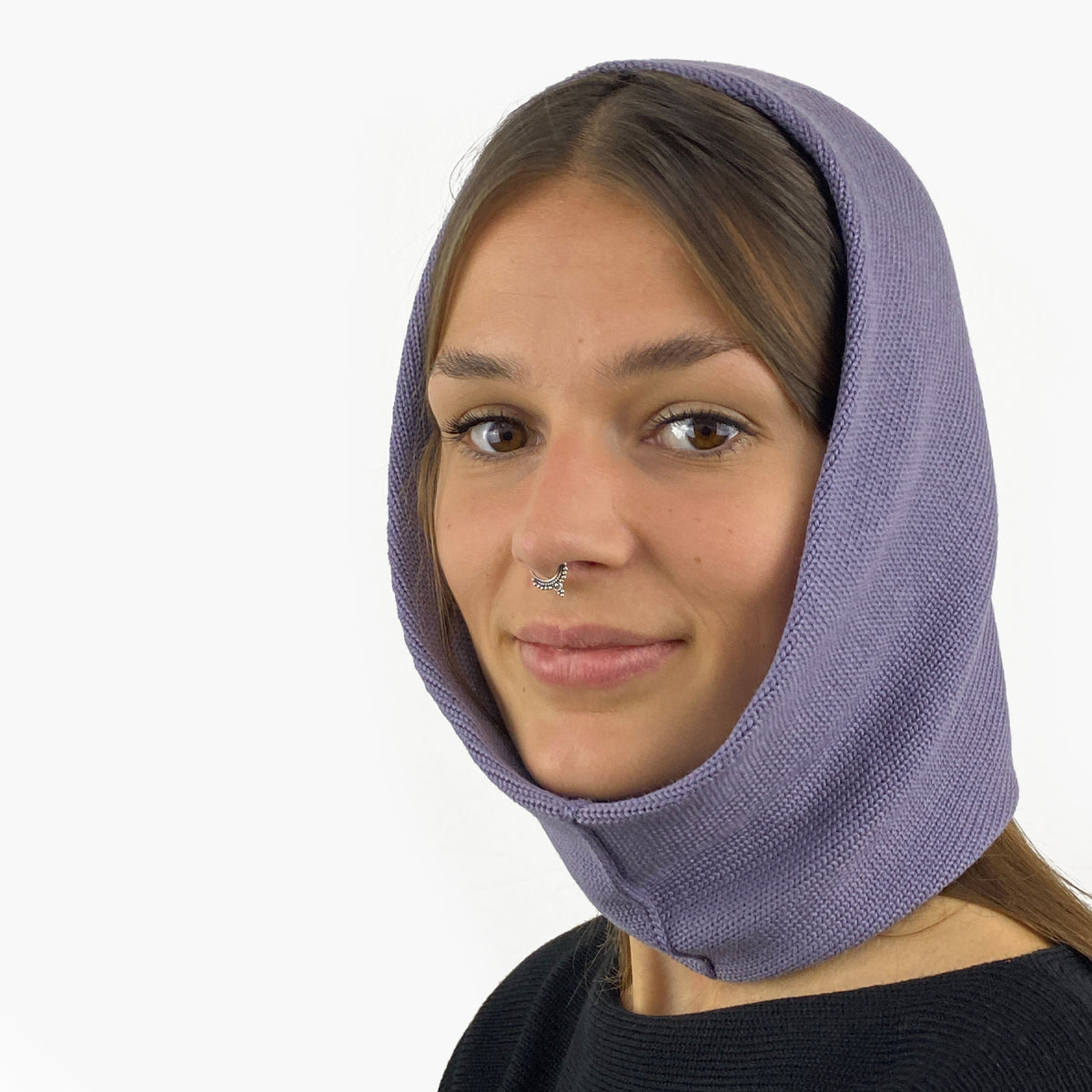 Eine junge Frau mit einem Nasenpiercing trägt eine Mütze deren Form den Kopf umspielt, und auch das Kinn bedeckt, ähnlich einer geschlossenen Kapuze. Die Balaclava ist Lilagrau und aus Merinowolle gestrickt.