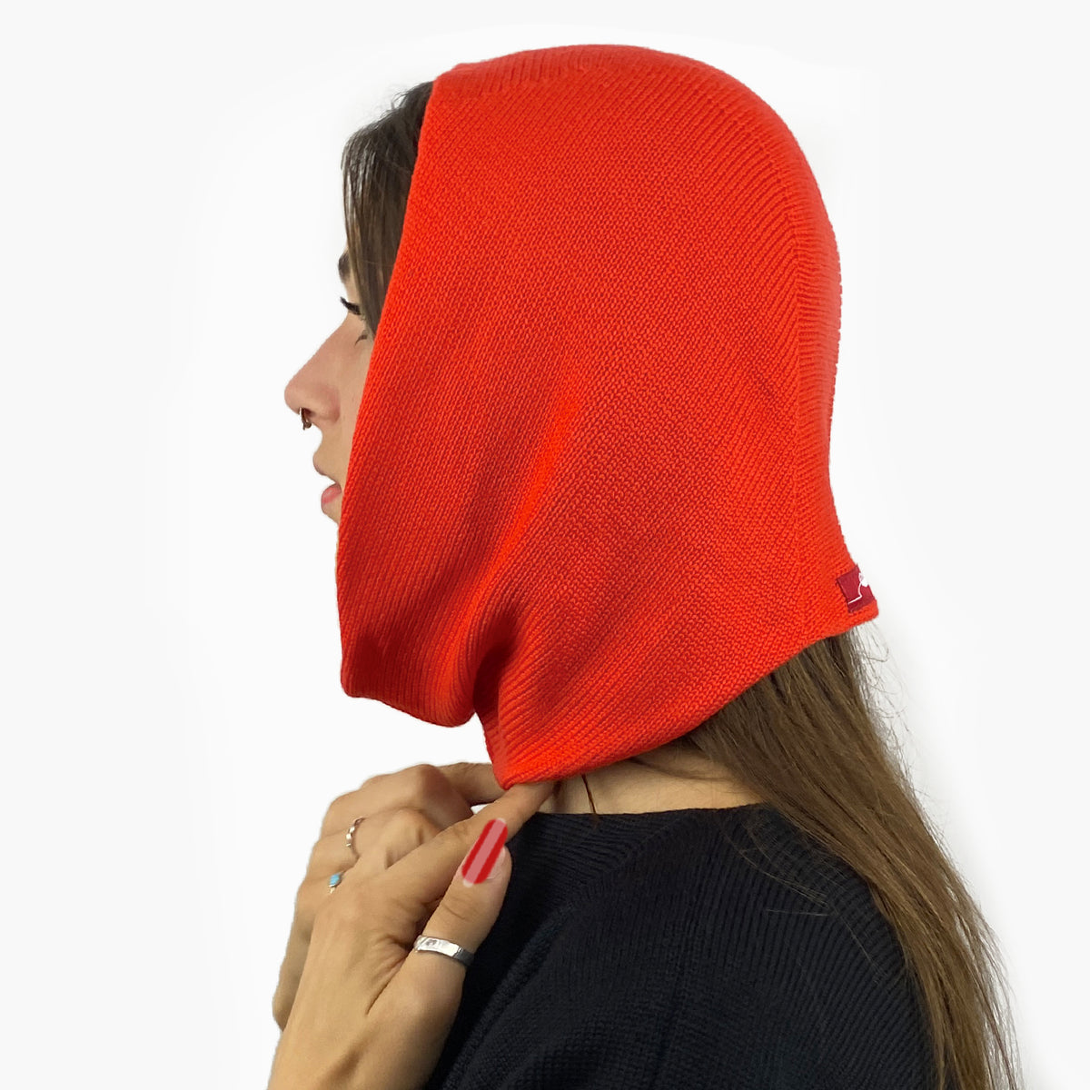 Eine junge Frau von der Seite zu sehen mit einem Nasenpiercing, trägt eine Mütze deren Form den Kopf umspielt, und auch das Kinn bedeckt, ähnlich einer geschlossenen Kapuze. Die Balaclava ist Orangerotund aus Merinowolle gestrickt.