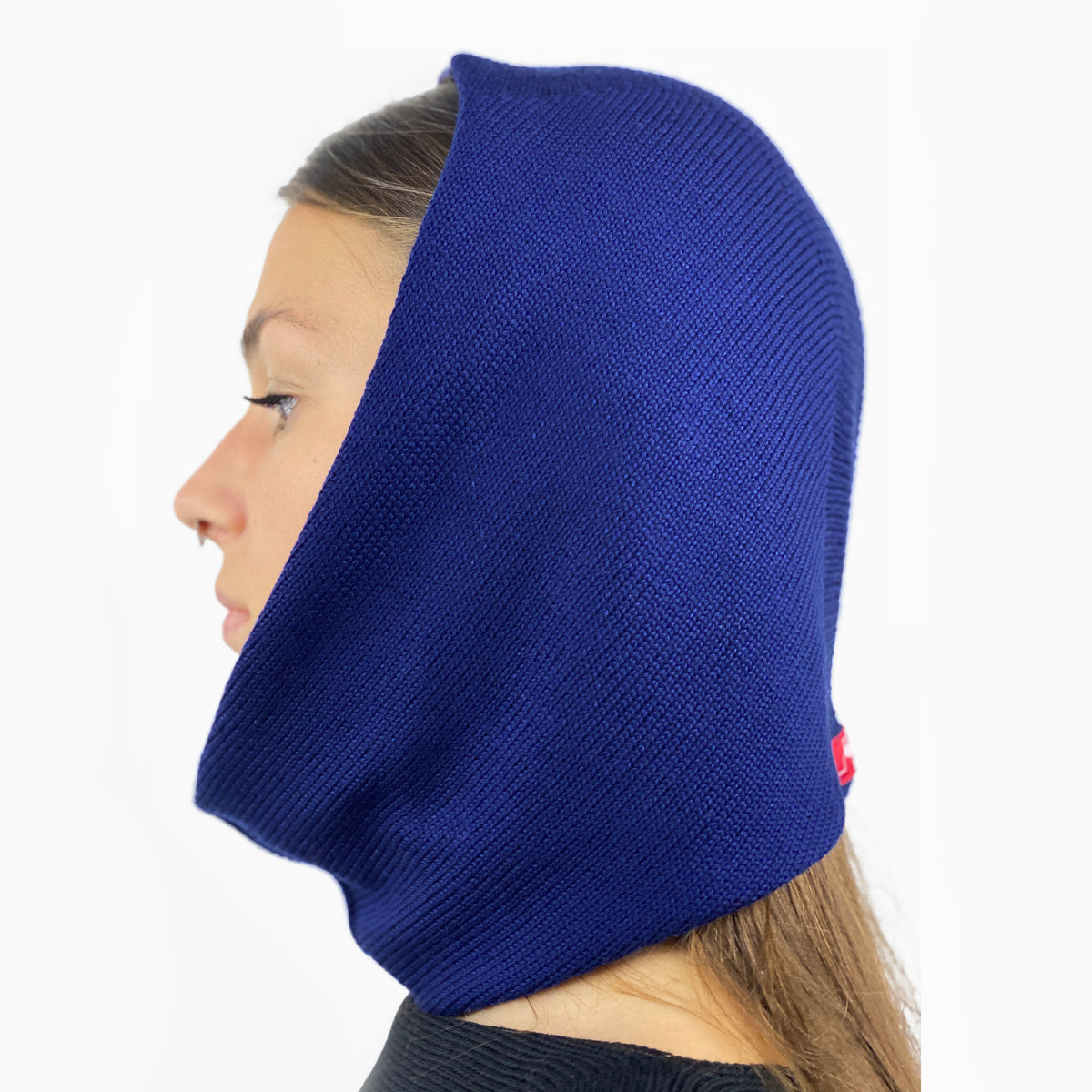 Eine junge Frau von der Seite zu sehen mit einem Nasenpiercing, trägt eine Mütze deren Form den Kopf umspielt, und auch das Kinn bedeckt, ähnlich einer geschlossenen Kapuze. Die Balaclava ist Royalblau und aus Merinowolle gestrickt.