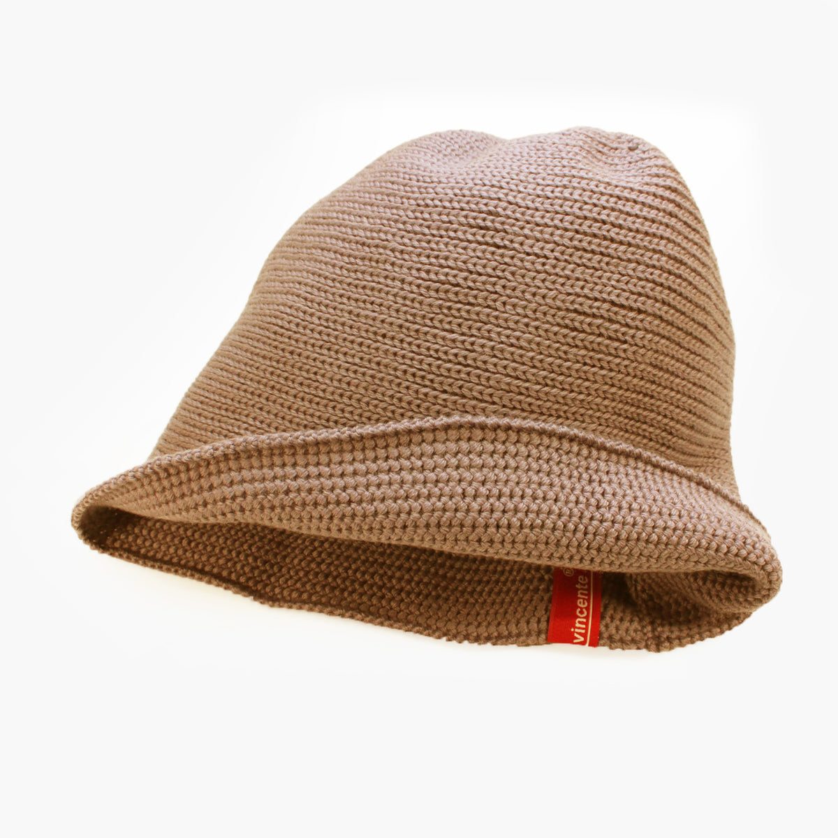 Karamel-Farbene, Beige Beanie-Mütze aus Merinowolle quer in Kopf-Form gestrickt. Das Fangpatenmuster bedeutet daß Innen ein Knötchenmuster und außen ein klasiches Rechte Maschenmuster zu sehen ist. Das rote vincente-Label mit der kleinen Ente ist am Rand angenäht. 