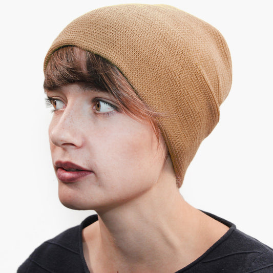 Eine Junge Frau trägt eine Karamel-Farbene, Beige Beanie-Mütze aus Merinowolle quer in Kopf-Form gestrickt. Das Fangpatenmuster bedeutet daß Innen ein Knötchenmuster und außen ein klasiches Rechte Maschenmuster zu sehen ist. Das rote vincente-Label mit der kleinen Ente ist am Rand angenäht. 