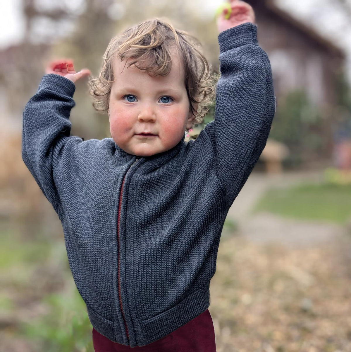 ein 2jähriges Mädchen streckt die arme nach oben. Sie trägt eine gestrickte Bomberjacke für Kinder mit Reißverschluß in grau.