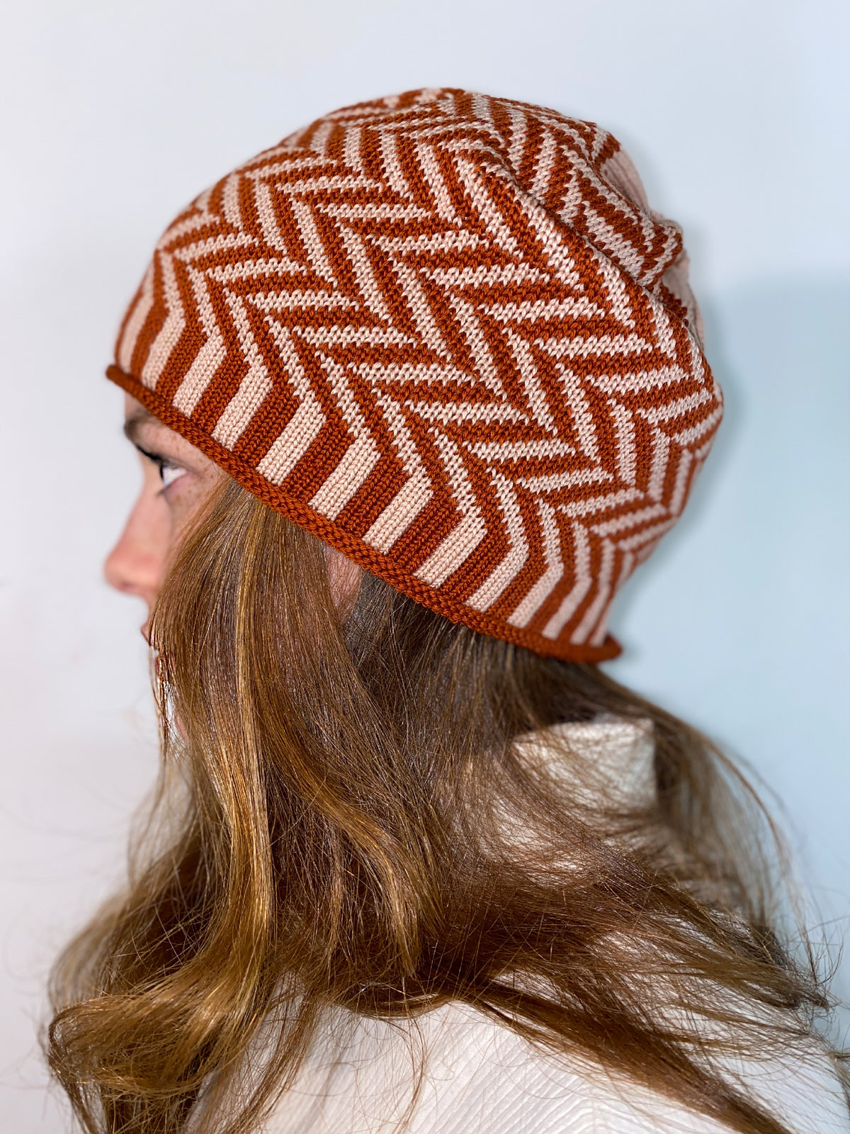 Eine Rothaarige junge Frau trägt eine Rostrote/Rosane Beanie-Mütze aus Merinowolle mit längs gestreiften elastischem Bündchen. Die Mütze ist im klassichem Fischgrätmuster gestrickt und hat eine kleine unauffällige Bommel.