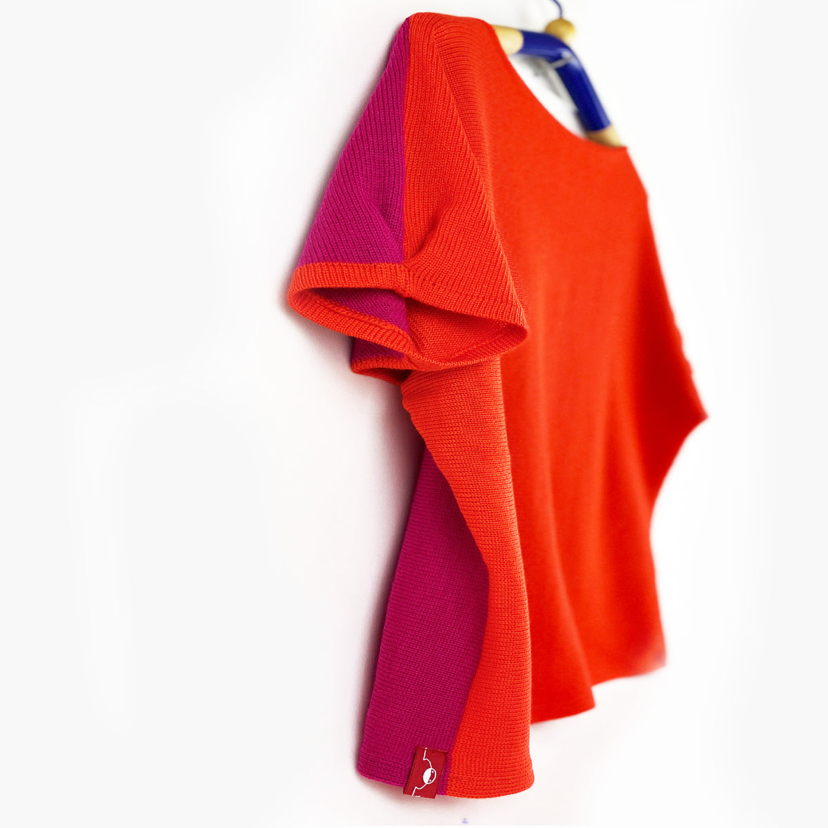 vorne Orangerot hinten Pink ist dieser quer gestrickterPullover mit kurzen, angeschnittenen Ärmeln ähnlich wie beim T-Shirt. Mit kleiner Falte am Armende. Man sieht das Kleidungsstück von der Seite auf einem Bügelhängend.