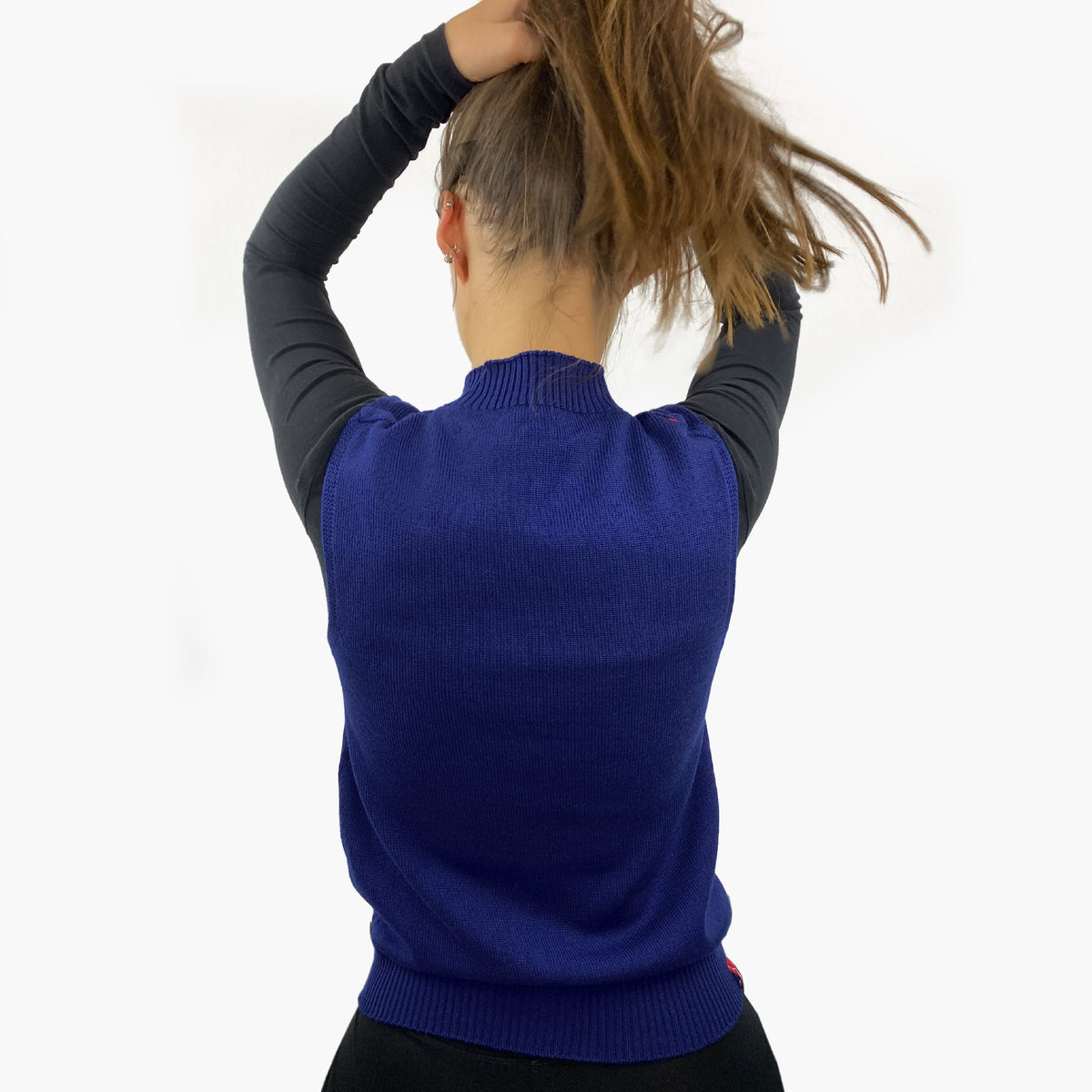 eine junge Frau von Hinten. Sie hält Ihre Haare nach oben. So sieht man den glatt gestrickten, Royalblauen Pullunder mit hochgeschnittenem Rundhalsauschnitt im rechts/links Muster. Auch der untere Abschluß ist im rechts/links Muster.