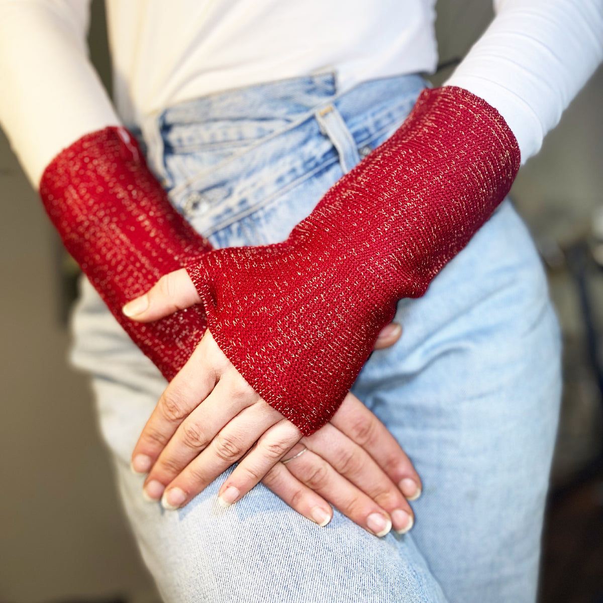 Zwei Hände überkreuzen sich im Schoß einer Jeans. Sie tragen Rote Armstulpen mit abgenähtem Daumenloch.Der Wolle ist ein Lurexfaden beigemischt. So daß die Stulpen weihnachtlich glitzern.