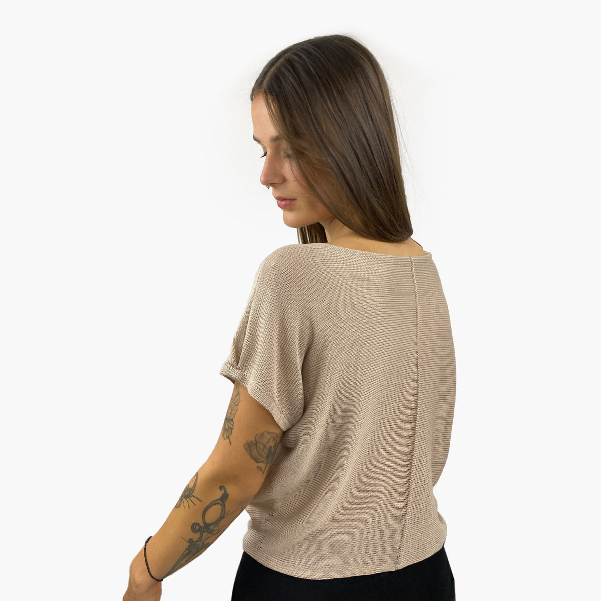 Unser Model mit glatten, Langem, braunem Haar und Taatoos (Schmetterling, Scorpion) auf dem Arm trägt ein seidig gänzendes T-shirt von vincente. Das Shirt ist aus Tencel/Lyocell gestrickt hat eine kleine Falte an dem kurzen Arm und ist Beige. Hier ist es von u hinten zu sehen, so daß man die Naht sieht, mit der das Shirt zusammengehäkelt wurde.