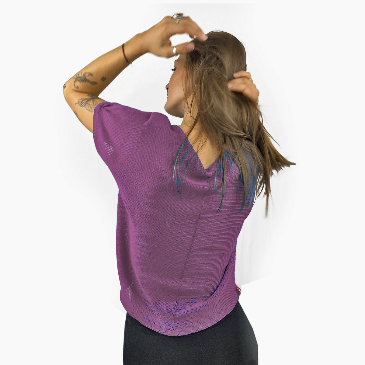 Unser Model mit glatten, Langem, braunem Haar und Taatoos (Schmetterling, Scorpion) auf dem Arm trägt ein seidig gänzendes T-shirt von vincente. Das Shirt ist aus Tencel/Lyocell gestrickt hat eine kleine Falte an dem kurzen Arm und ist Lila. Hier ist es von hinten zu sehen, so daß man die Naht sieht, mit der das Shirt zusammengehäkelt wurde.