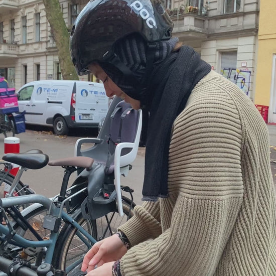 Film in dem eine junge Frau den Schal mit eingearbeiteter Kapuze im Fischgraetmuster unter ihrem Fahrradhelm trägt. Der Schal ist Grau und Schwarz.