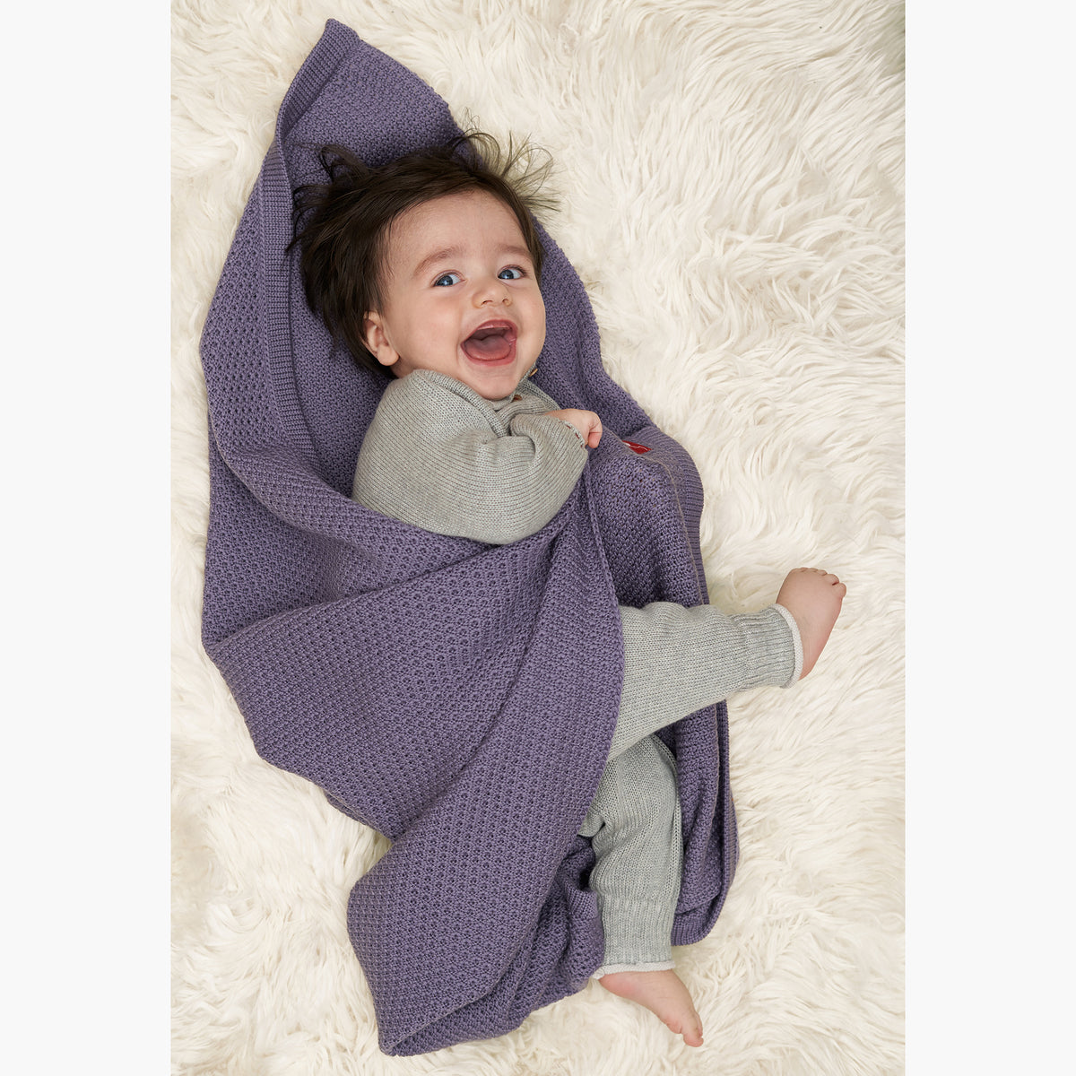 Ein kleines Baby lacht mit weit aufgesissem Mund und Augen. Es hat noch keine Zähne aber extrem viele schwarze Haare. Das baby ist in eine Lilane im Fangpatentmuster gestrickte Babydecke von vincente gehüllt