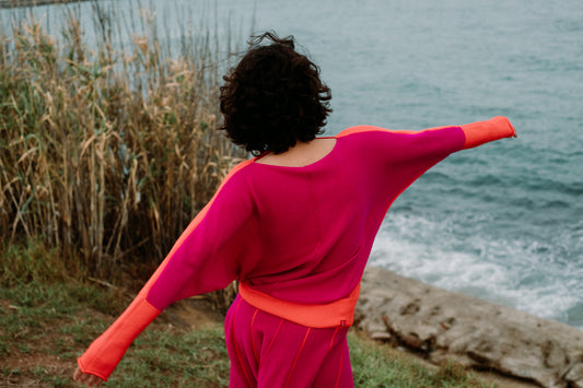 Ein langärmeliger Pullover in leuchtendem Fuchsia-Pink mit hellorangen Akzenten an den Ärmeln und am Saum. Das Kleidungsstück hat einen weiten Ausschnitt und eine lockere Passform mit verlängerten Ärmeln, die über die Handgelenke reichen, und zeigt ein kräftiges Farbblock-Design.