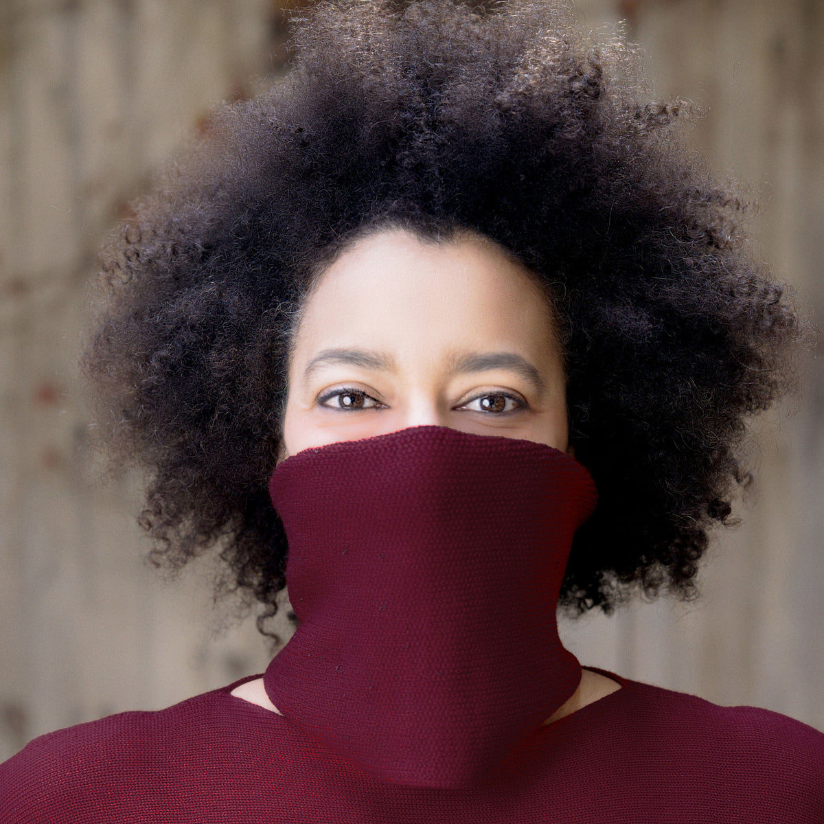 eine junge Frau mit großen Braunen Augen und Langen braunen Afro, trägt einen Bordeaux farbenen Schlauchschal um den Hals und verdeckt auch Ihr halbes Gesicht, so daß die Augen besonders stralen.