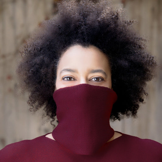 eine junge Frau mit großen Braunen Augen und Langen braunen Afro, trägt einen Bordeaux farbenen Schlauchschal um den Hals und verdeckt auch Ihr halbes Gesicht, so daß die Augen besonders stralen.