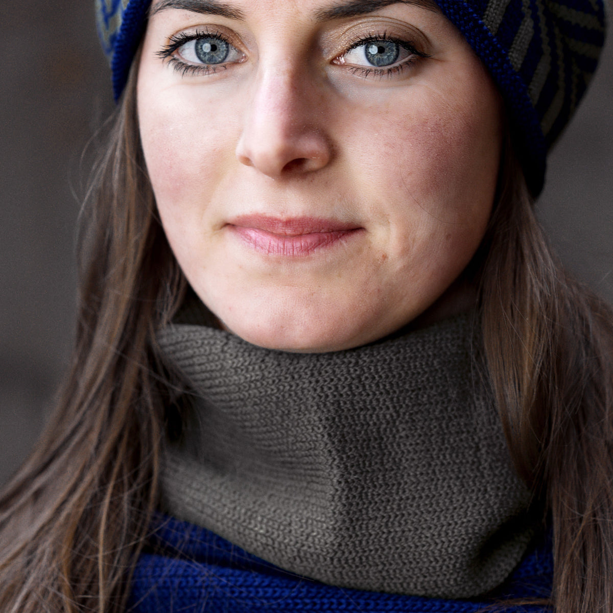 eine junge Frau mit großen blauen Augen und Langen braunen Haaren trägt einen Grauen Schlauchschal um den Hals.