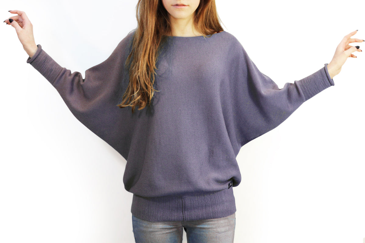 ein Modell mit schwarzen langen Fingernägeln breitet die Arme aus um den Schnitt des lilanen Fledermausullover zu präsentieren. Der gestrickte Pullover hat einen elastichen Bund und lange elastische Ärmelbündchen.