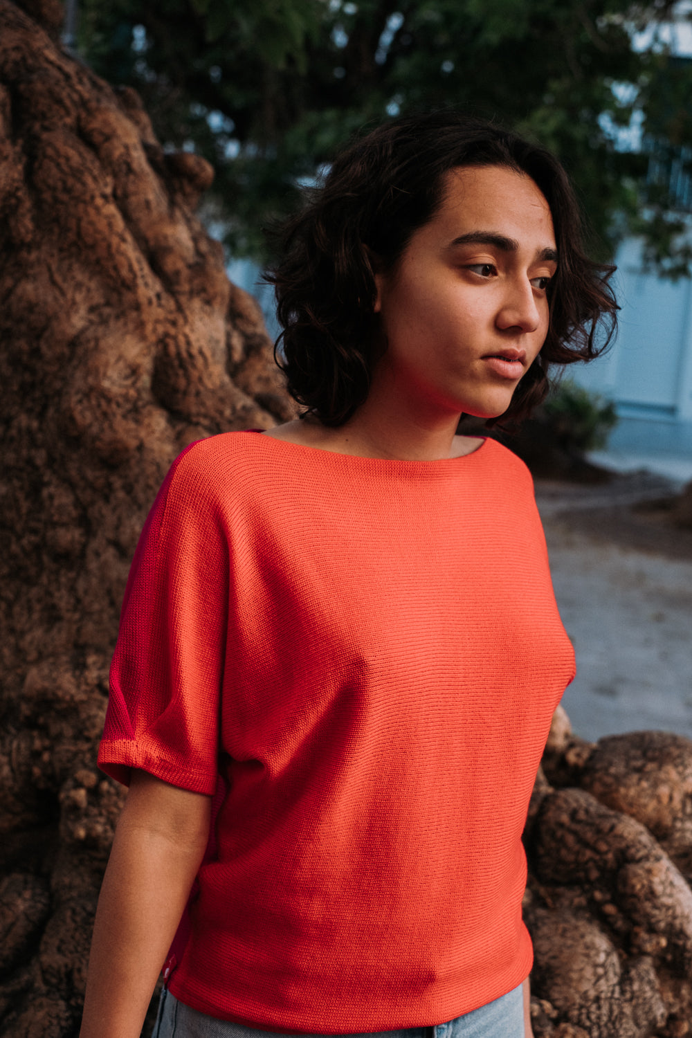 Eine junge Frau mit schwarzen Locken steht unter einem alten Olivenbaum. Sie trägt einen kurzärmeligen Pullover von vincente. Das T-shirt ähnliche Bekeidungstück hat eine Falte am Arm und ist aus Orangeroter Merinowolle gestrickt.