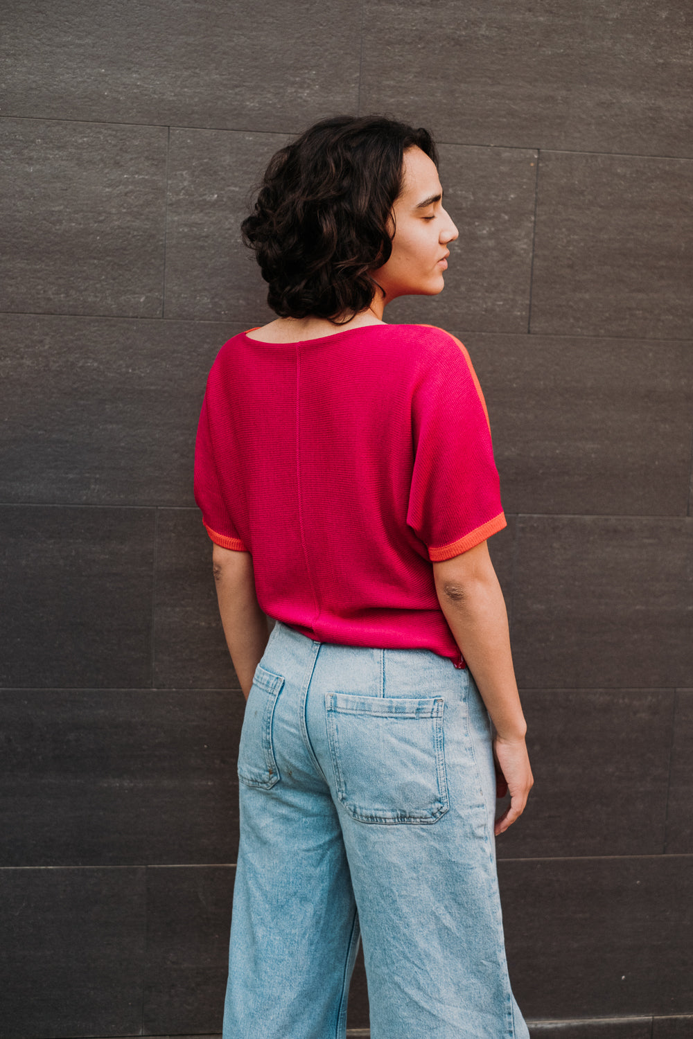 Eine junge Frau mit schwarzen Locken steht vor einer Grauen Steinwand. Sie trägt einen kurzärmeligen Pullover von vincente. Das T-shirt ähnliche Bekeidungstück hat eine Falte am Arm und ist vorne aus Orangeroter Merinowolle und Hinten in Pink gestrickt.