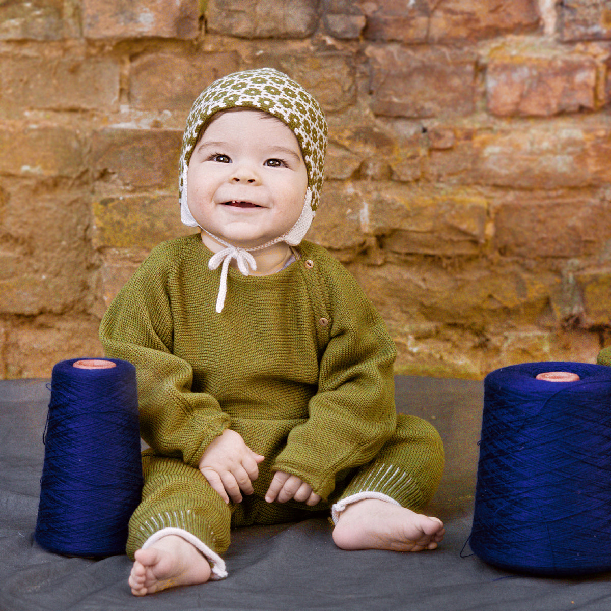Lachendes Baby trägt eine gepunktete Mütze und eine Olivegrüne Babyjacke mit seitlicher Knöpfung, auch Schlüttli genannt. Die Strickjacke ist aus Bio-Merinowolle gestrickt