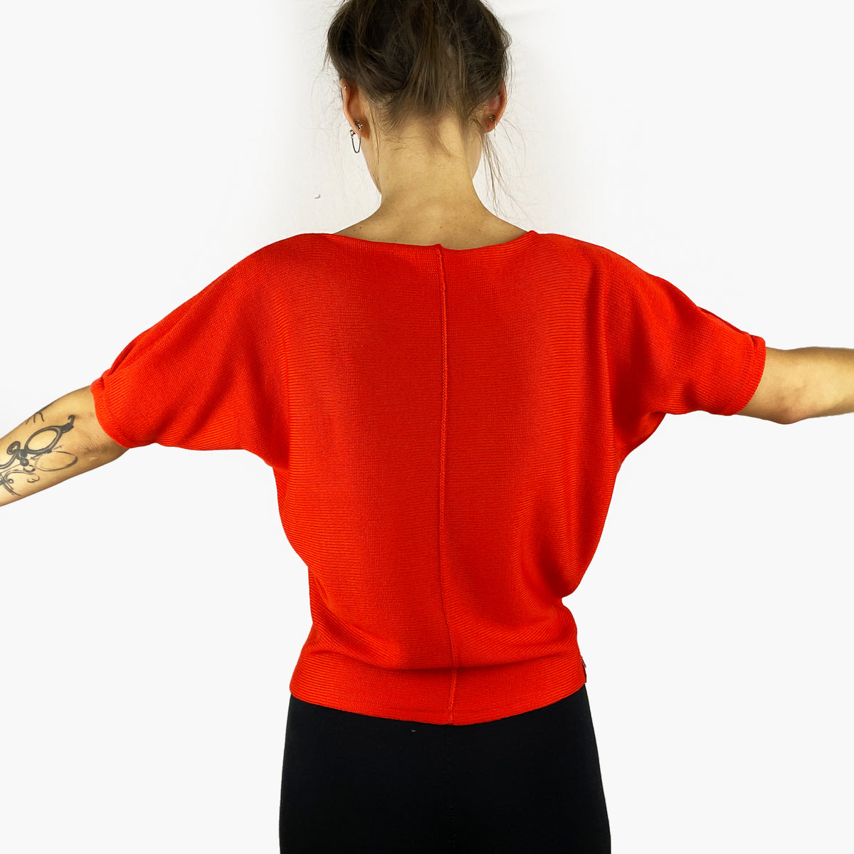 Man sieht ein Model von Hinten. es trägt einen Orangeroten gestrickten Pullover mit kurzen, angeschnittenen Ärmeln ähnlich wie beieinem  T-Shirt. Mit kleiner Falte am Armende. In der hinteren Mitte, die man hier sieht ist eine längsverlaufende Häkelnaht.
