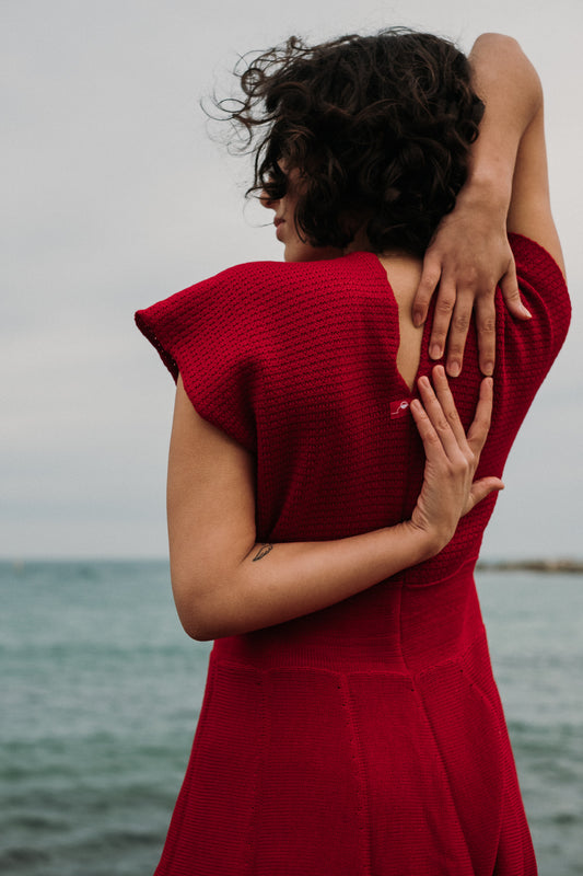 Eine junge Frau mit schwarzen Locken steht am Strand, im Hintergrund das Meer. Man sieht Sie von Hinten, so daß man den V-ausschnitt am Rücken sieht. Es ist ein Rotes schwingendes Kleid mit angeschnittenen Ärmeln aus feiner Bio-Merinowolle gestrickt von vincente