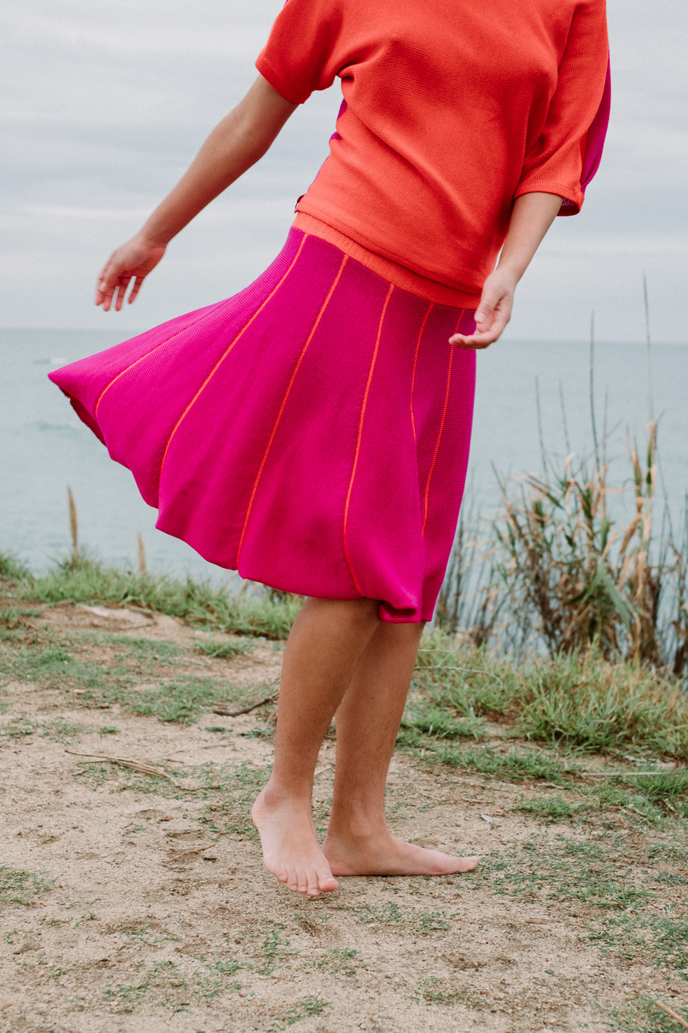 Ein junges Mädchen mit schwarzen Locken  schwingt Ihren Rock am Strand. Der Rock ist Pink und hat Orange Längsstreifen. Er ist aus Merinowolle gesrtrickt. Auch das Oberteil von vincente ist in den 2 Farben gestrickt.
