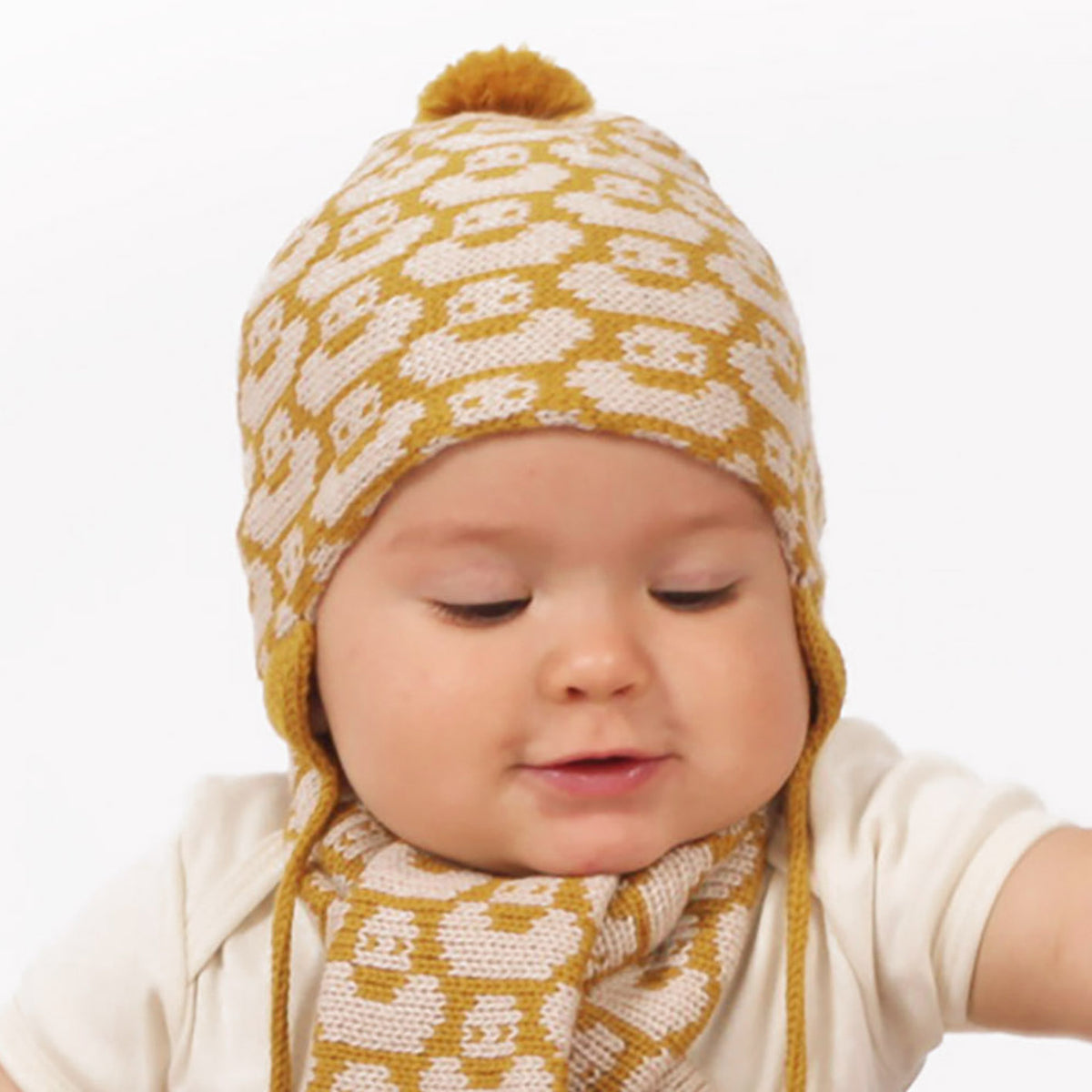 Ein kleines Baby guckt neugierig in die Welt. Es trägt eine Gold/Wollweiße Mütze mit einem fortlaufenden Muster mit kleinen Enten. Die Babymütze hat eine Bommel und angestrickte Ohrläppchen mit Bändern zum festbinden. Sie ist aufwendig der Kopfform nachgestrickt und paßt sich sehr gut an. Das rote Label mit der kleinen Ente ist hinten angenäht.