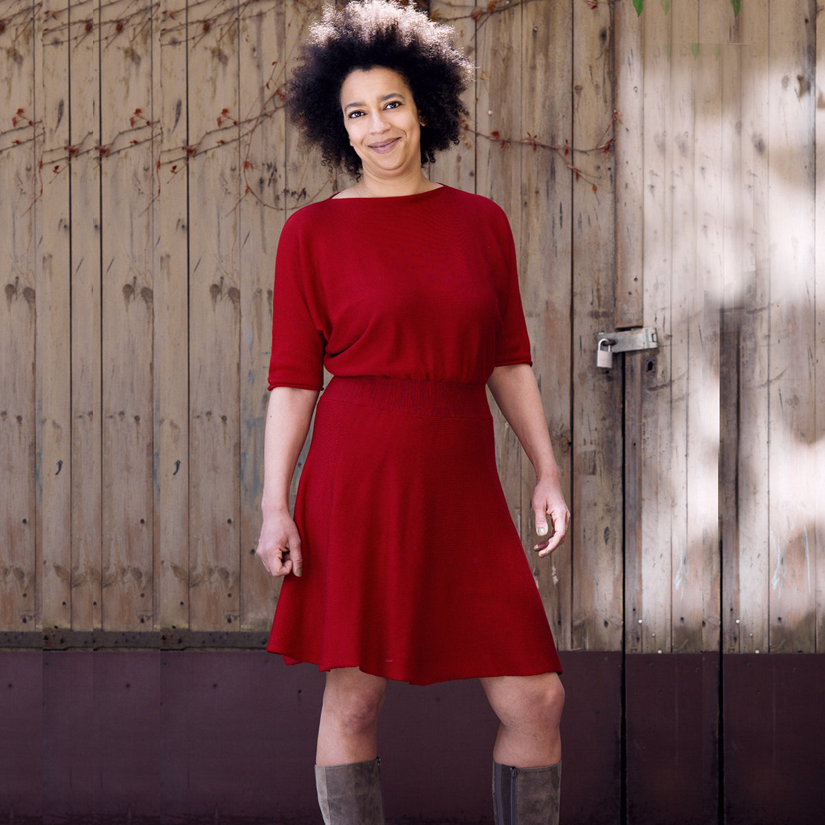 Eine Junge Frau mit Afrohaaren trägt ein Rotes, tailliertes Strickkleid mit Fledermausärmeln 3/4lang und ausgestellten Rock. Sie steht vor einem hölzernen Scheunentor.