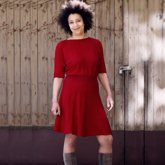 Eine Junge Frau mit Afrohaaren trägt ein Rotes, tailliertes Strickkleid mit Fledermausärmeln 3/4lang und ausgestellten Rock. Sie steht vor einem hölzernen Scheunentor.