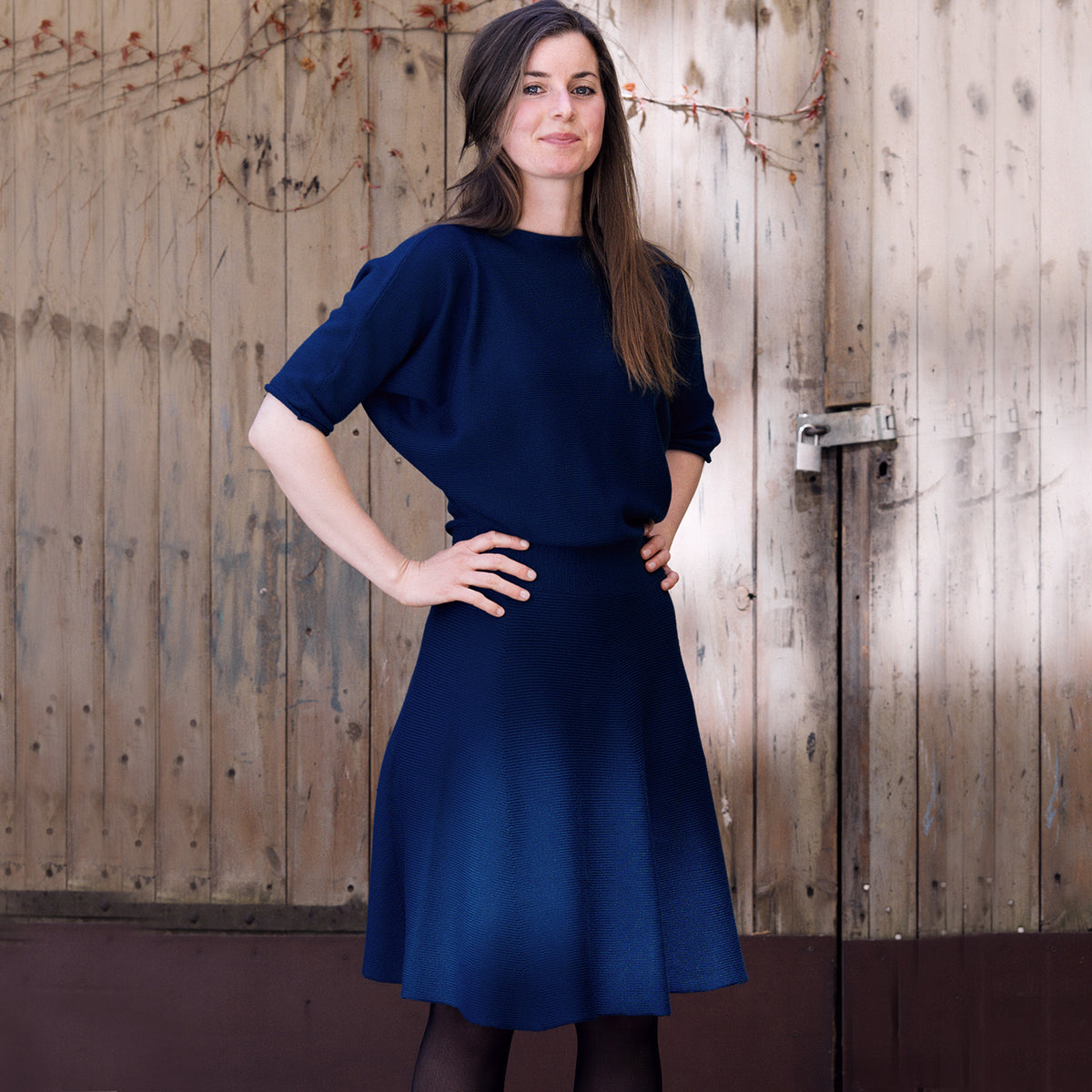 Eine Junge Frau mit langen braunenHaaren trägt ein Royalblaues, tailliertes Strickkleid mit 3/4langen Fledermausärmeln und ausgestellten Rock. Sie steht vor einem hölzernen Scheunentor.