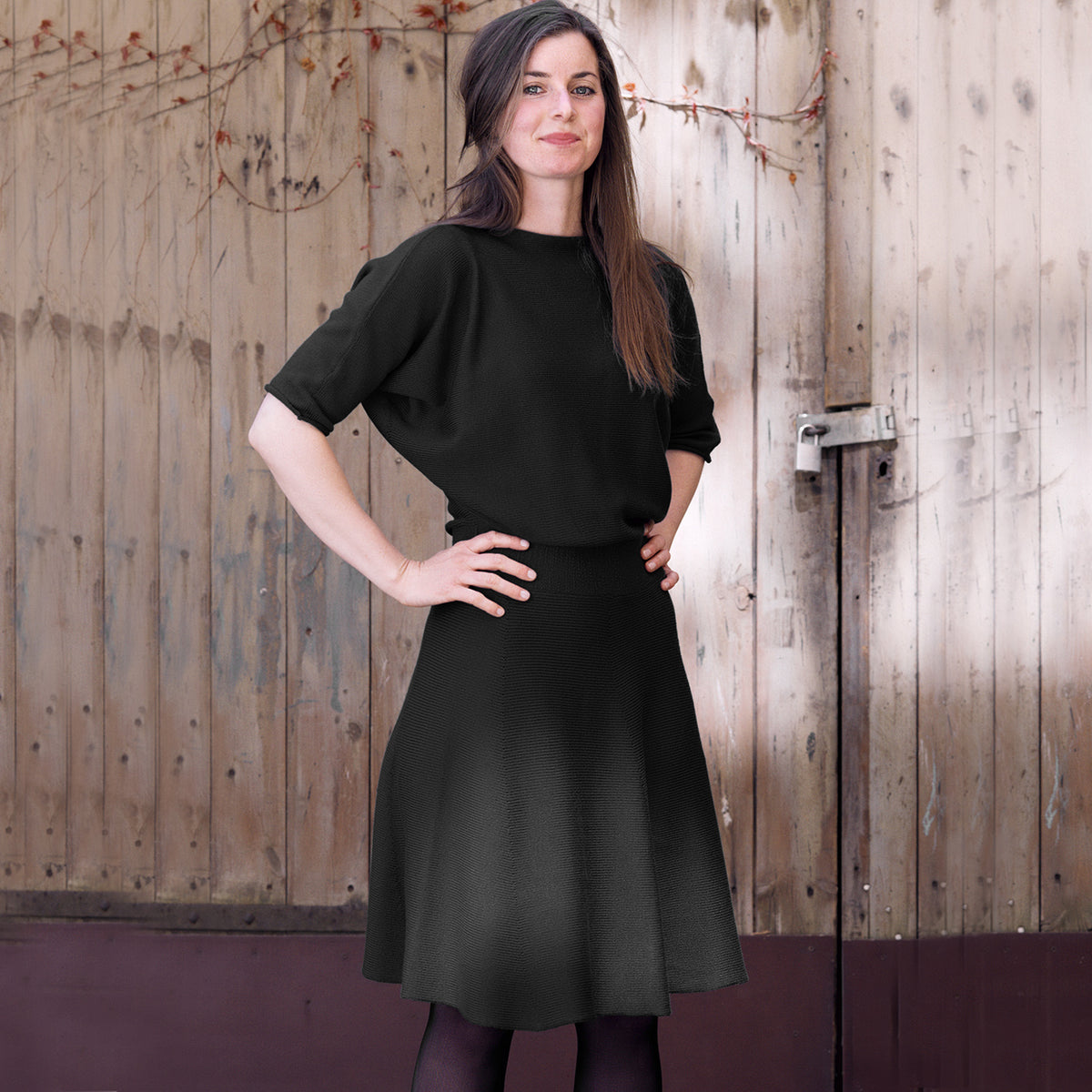 Eine Junge Frau  trägt ein schwarzes, tailliertes Strickkleid mit 3/4langen Fledermausärmeln und ausgestellten Rock. Handgefertigt aus feinster zertifizierter Merinowolle aus dem Atelier vincente