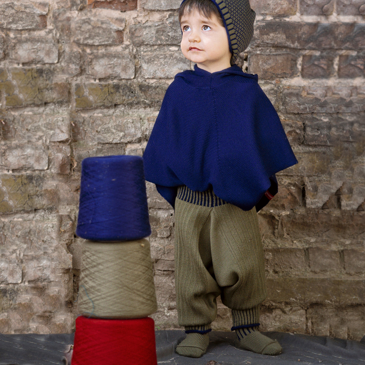 Ein kleiner Junge hat einen Turm aud Wollkonen gebaut. Er trägt einen Royalblauen Poncho und eine Olivegrüne Sarouelhose von vincente