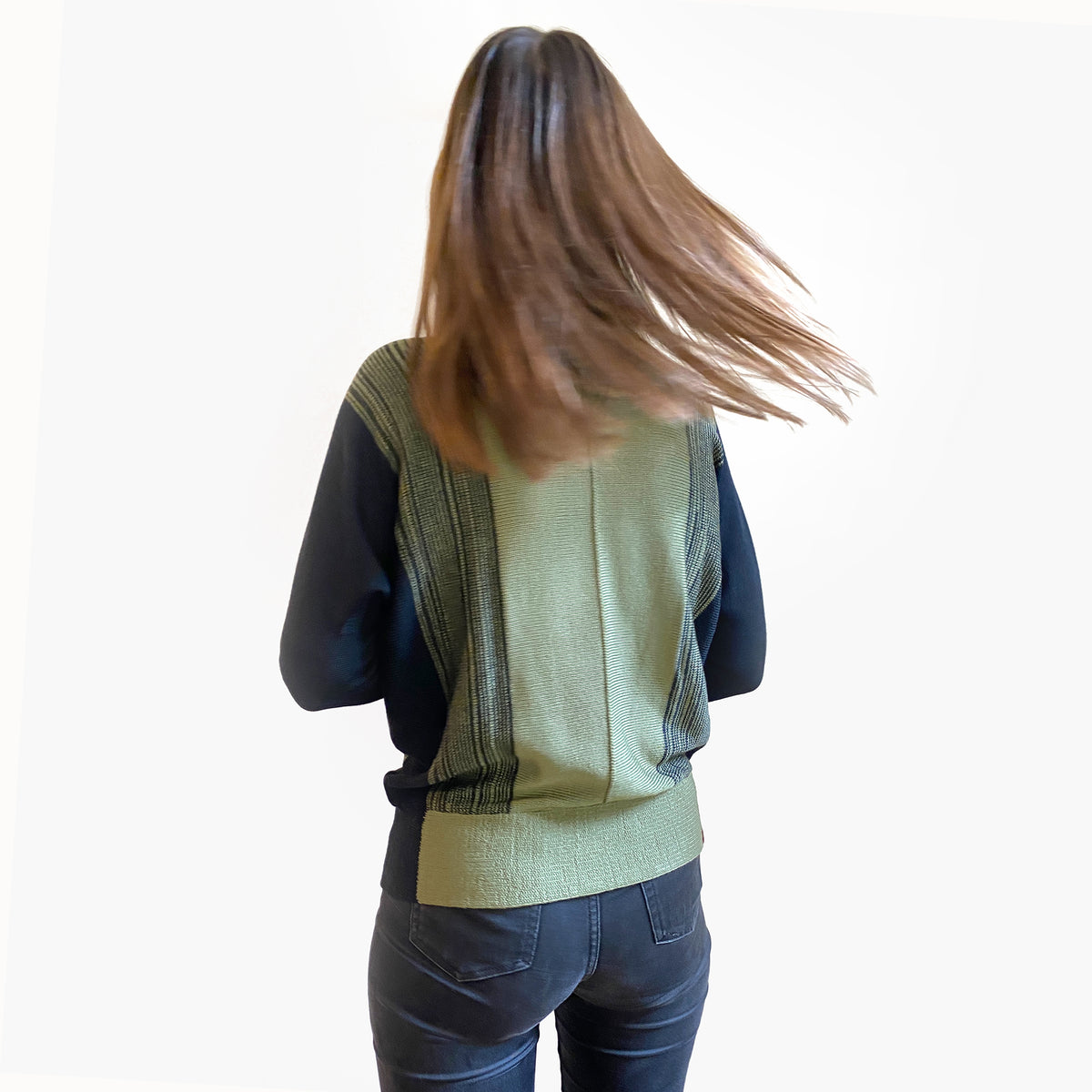 Man sieht ein Modell von Hinten, mit braunen langen Haaren. Sie trägt einen gestrickten Wollpullover mit Farbverlauf von Olive nach Schwarz