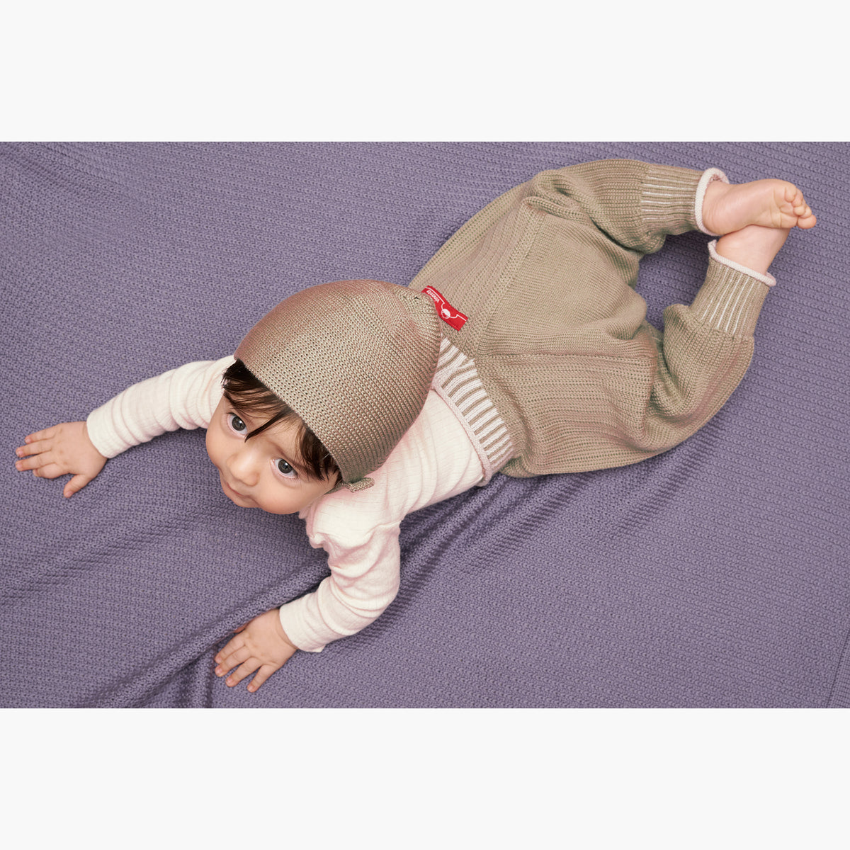 ein Baby mit einer karamell-farbenen Haube liegt auf einer Lila farbenen Babydecke und trägt eine Sarouelhose auch Harems- oder Pluderhose genannt. Mit tiefgeschnittenem Schritt und extrem viel Platz für Windeln. Die Karamell-Farbene  Wollhose ist aus feinster Bio-Merinowolle gestrickt. Sie hat enganliegende hochelastischen Bündchen, die Wollweiße Längsstreifen haben.