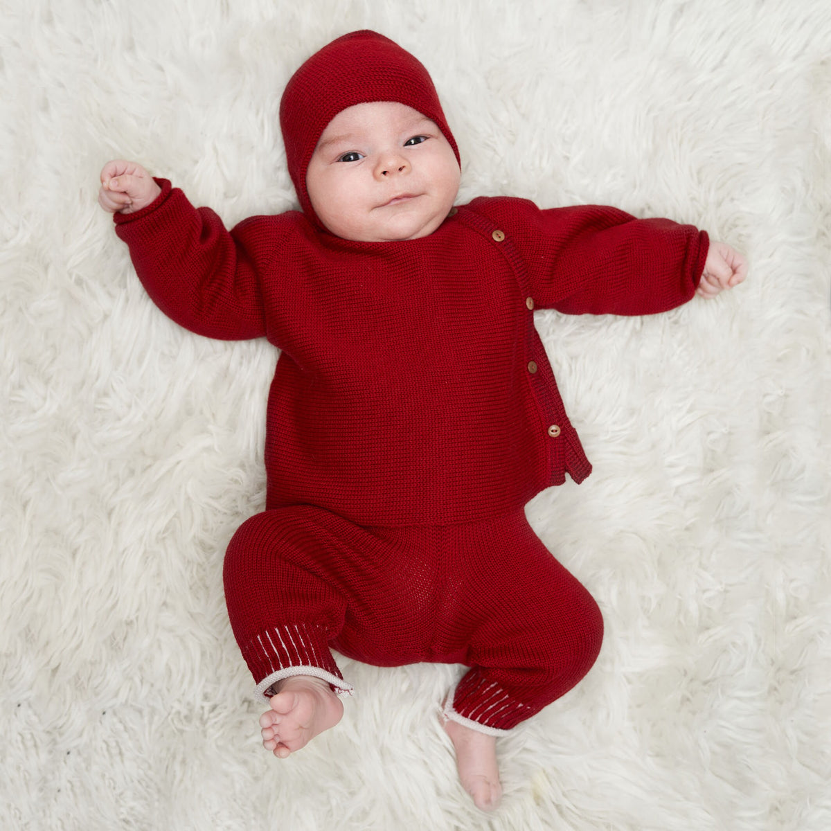 Baby trägt eine rote Haube und eine Rote Babyjacke mit seitlicher Knöpfung, auch Schlüttli genannt. Die Strickjacke ist aus Bio-Merinowolle gestrickt. Dazu eine rote Salopette-Hose von vincente