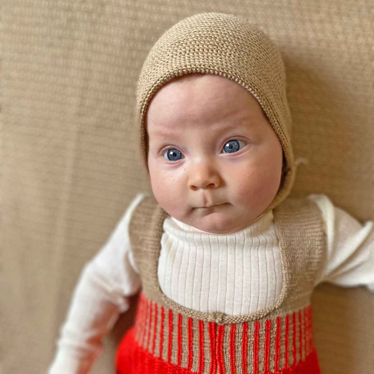 Pausbäckiges Baby guckt mit großen blauen Augen in die Welt hinaus. Es trägt eine beige gestrickte Haube von vincente. Mit verstellbarem Bändchen zum zuknöpfen und kleinem beigen Knopf.