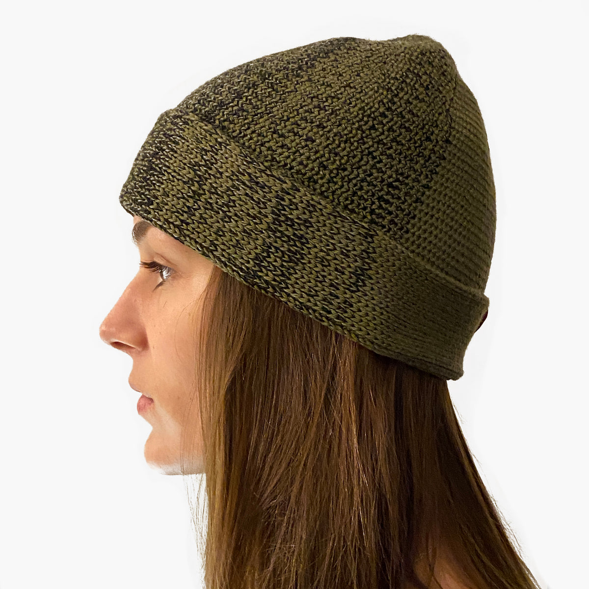 Eine Junge Frau trägt eine Olive/Schwarze  Beanie. Der Rand ist umgeschlagen, wie bei einer Dockermütze.  Die Mütze hat einen Farbverlauf, der sich im Kreis wie Tortenstücke verhält. Ein Stück ist Olive-farben ein Stück Schwarz. Dazwischen  michen sich die beiden Farben im Farbverlauf von Olive mit etwas Schwarz dann etwas mehr Schwarz usw.