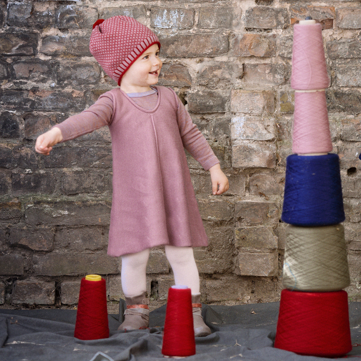 Ein kleines Mdchen freut sich über den Turm aus 5 bunten Wollkonen, den es gebaut hat. Es ist in einem rosanen Strickkleid in A-Form vin vincente gekleidet
