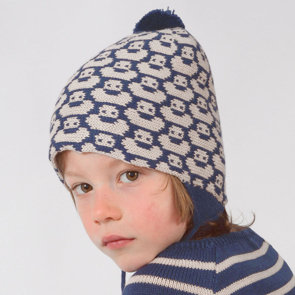 ein 4jähriger Junge trägt eine Blau/Wollweiße mit einem fortlaufenden Muster mit kleinen Enten. Die Babymütze hat eine Bommel und angestrickte Ohrläppchen mit Bändern zum festbinden. Sie ist aufwendig der Kopfform nachgestrickt und paßt sich sehr gut an. Das rote Label mit der kleinen Ente ist hinten angenäht.