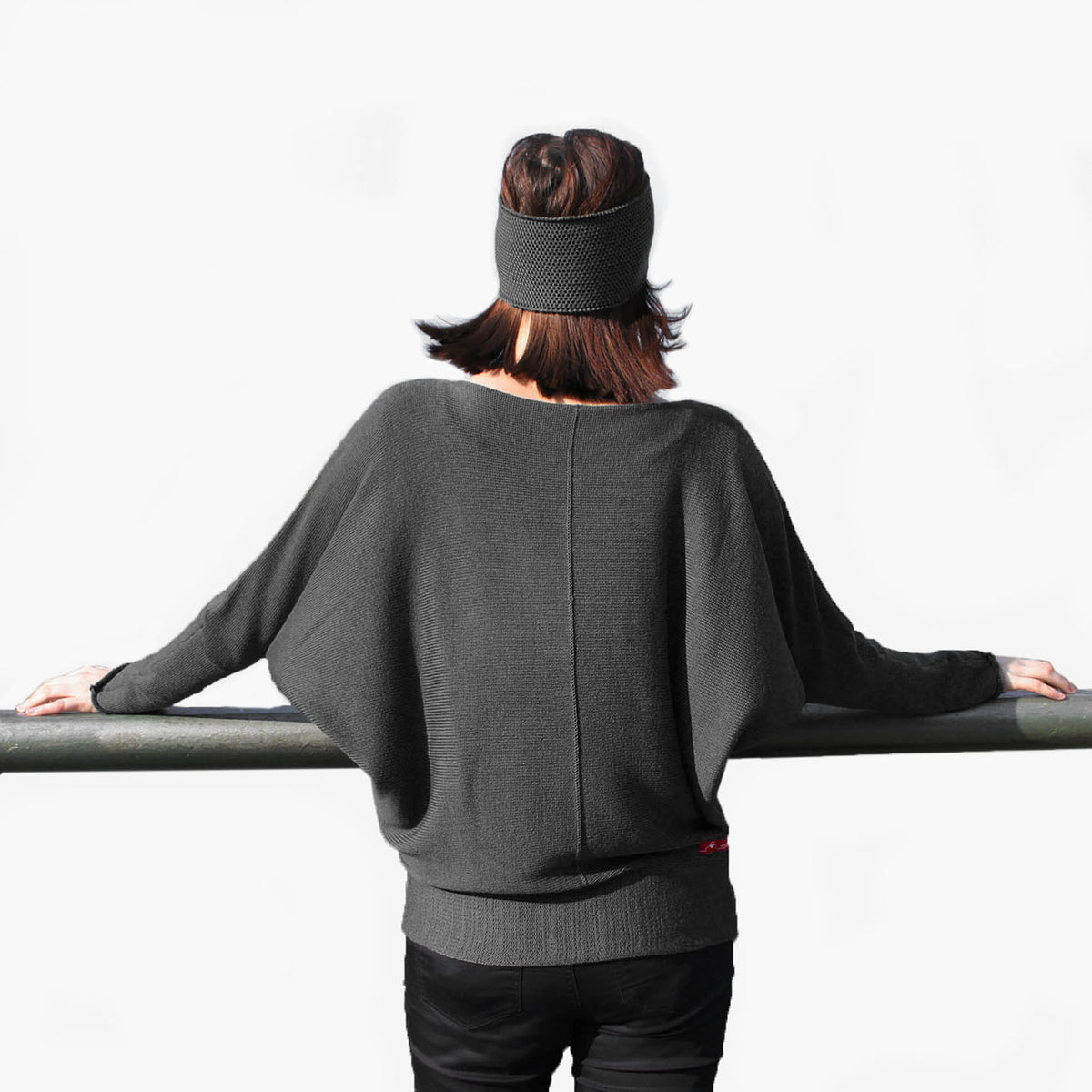 Man sieht eine junge Frau von Hinten. Sie trägt ein graues Stirnband. Sie breitet die Arme aus um den Schnitt des grauen Fledermausullover zu präsentieren. Der gestrickte Pullover hat einen elastichen Bund und lange elastische Ärmelbündchen.