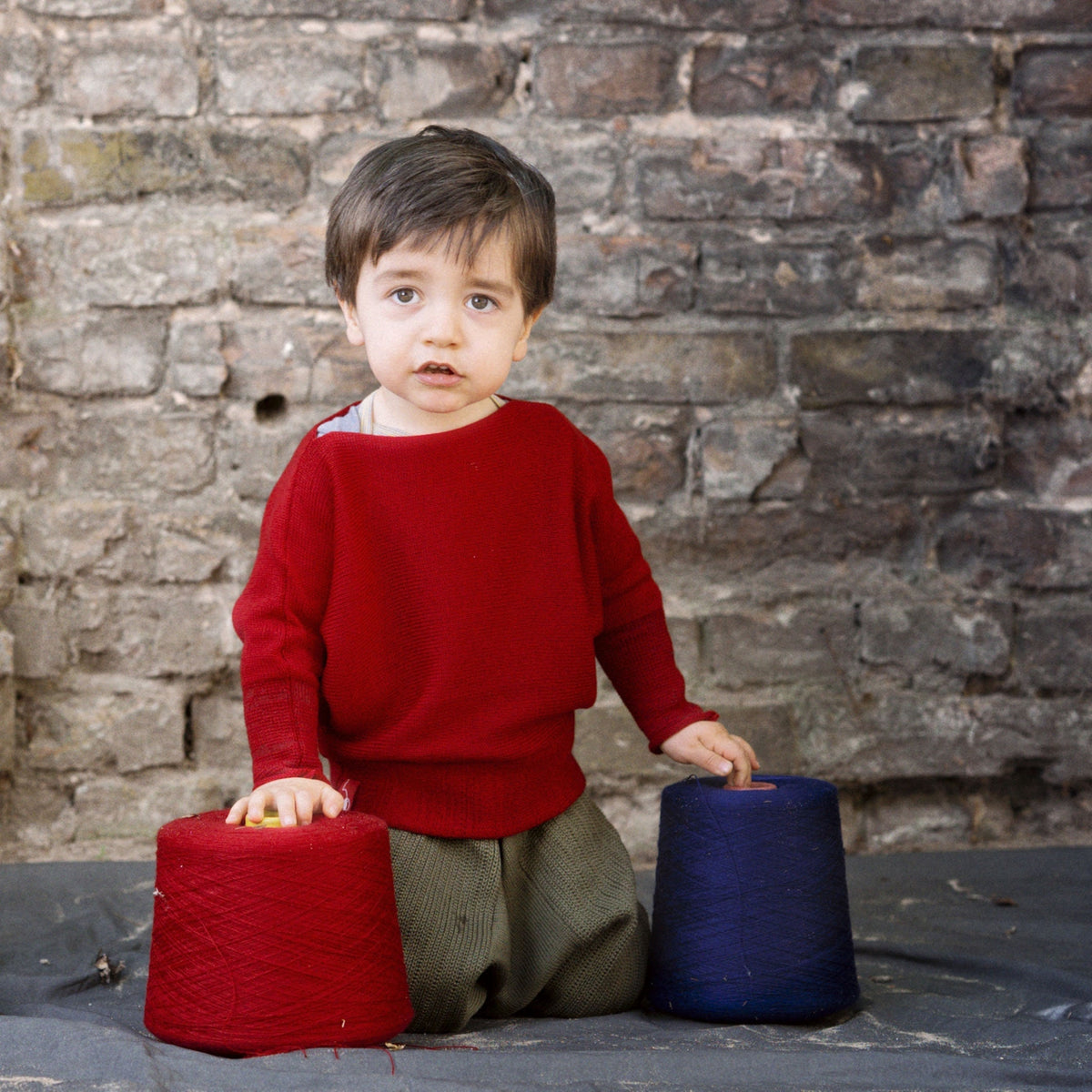 Ein kleiner Junge spielt mit Wollkonen. Er trägt einen Roten gestrickten Pullover mit angeschnittenen Ärmeln. Der Pullover hat einen elastichen Bund und lange elastische Ärmelbündchen.